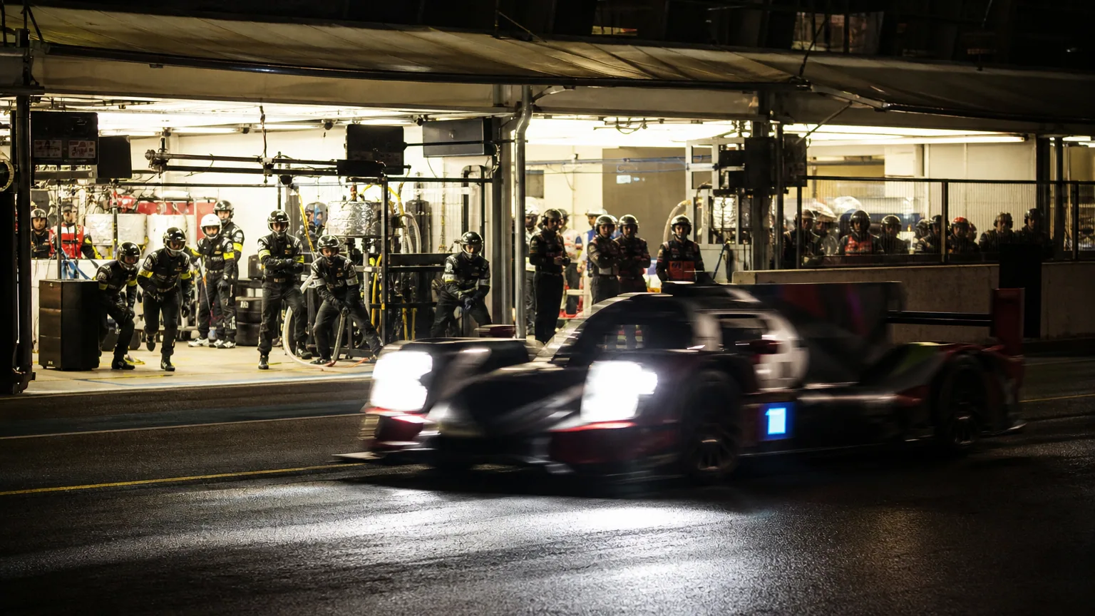 Voiture de course prototype lancée à pleine vitesse de nuit sur le circuit, stands éclairés en arrière-plan