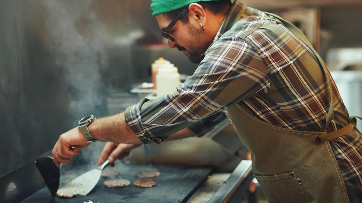 Un cuisinier saisit des steaks hachés à la spatule sur une plaque de cuisson brûlante