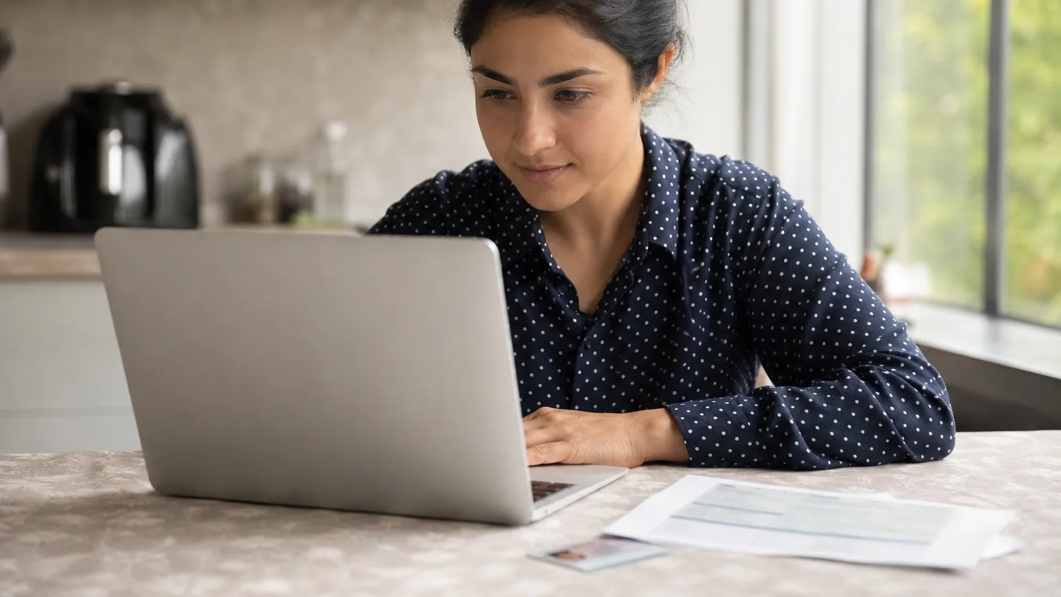 Une femme remplit son inscription en ligne sur un ordinateur portable avec ses documents administratifs posés à côté