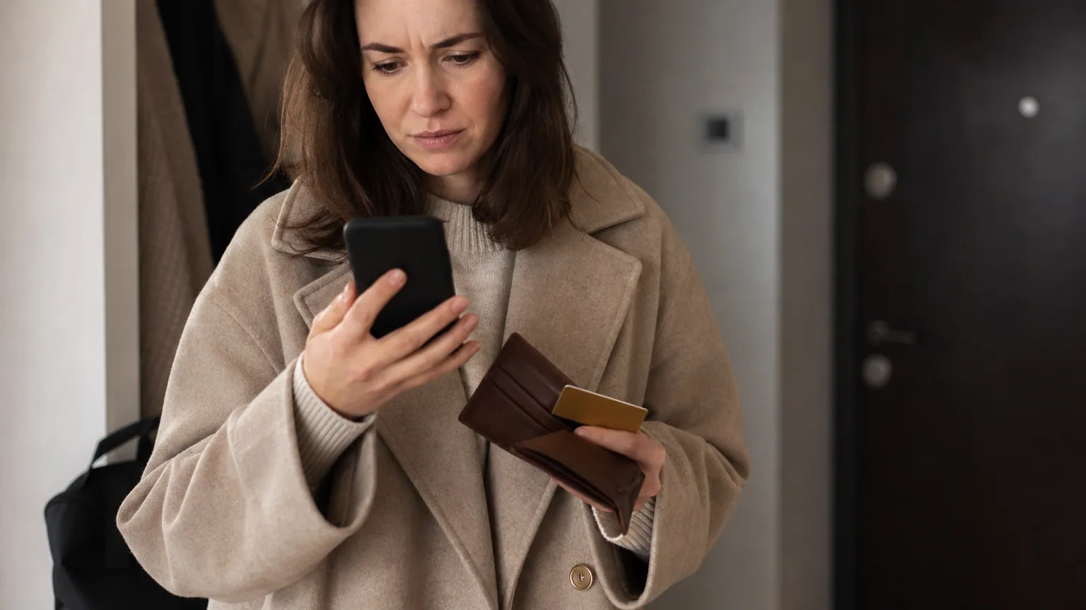 Une personne compose un numéro sur son téléphone, carte bancaire à la main, l'air préoccupé dans une entrée d'immeuble
