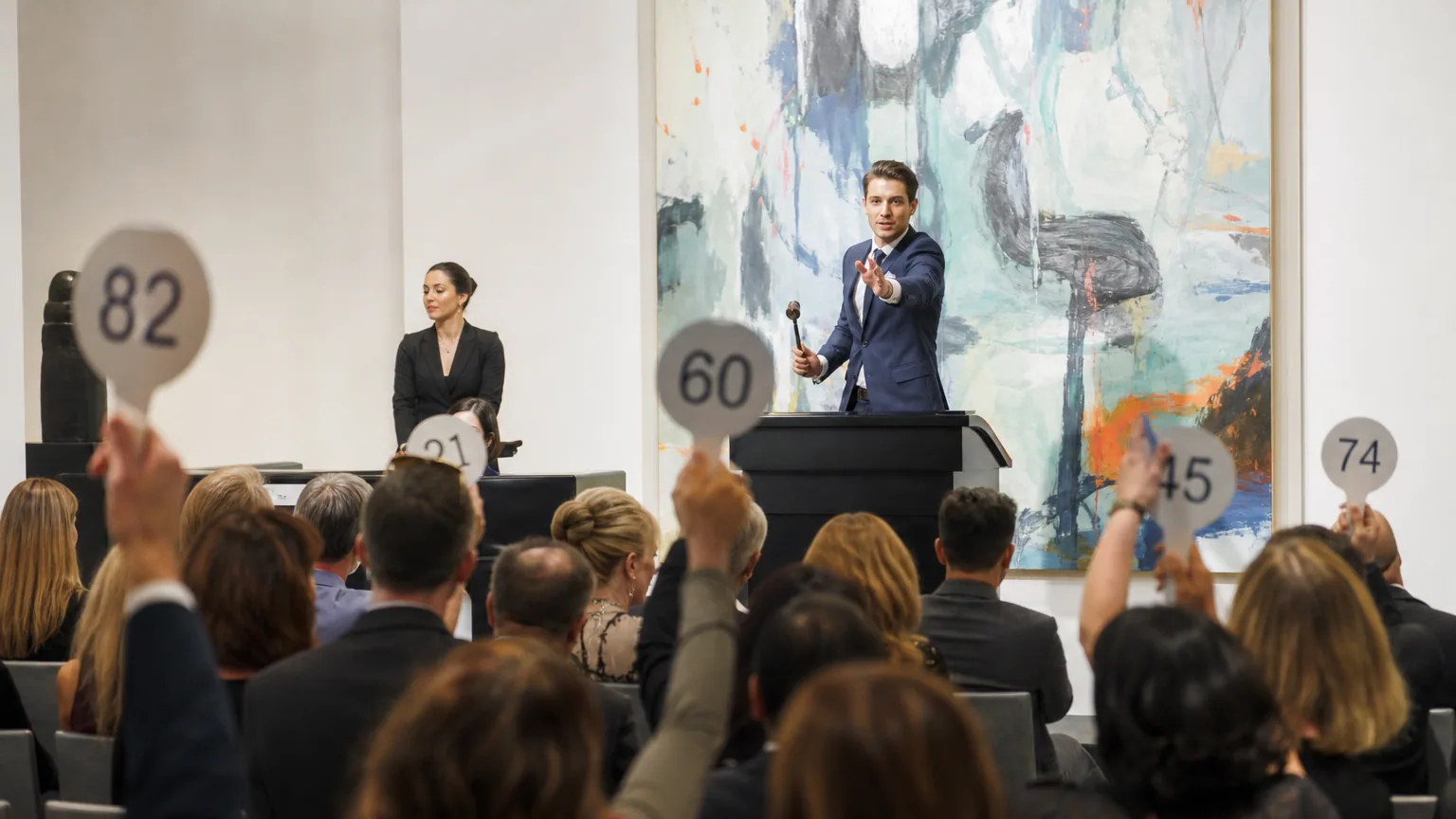 Salle de vente aux enchères d'art contemporain avec un commissaire-priseur et des collectionneurs levant leurs palettes