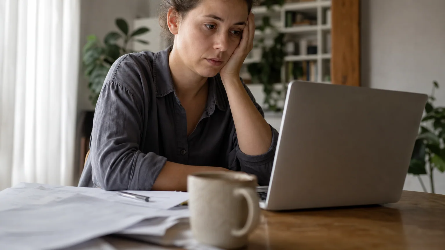 Une personne assise à son bureau à la maison, hésitante devant son ordinateur portable et des documents