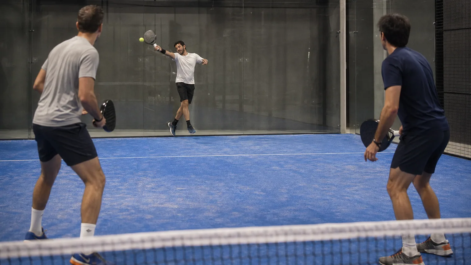 Quatre joueurs en plein échange sur un terrain de padel entouré de parois vitrées