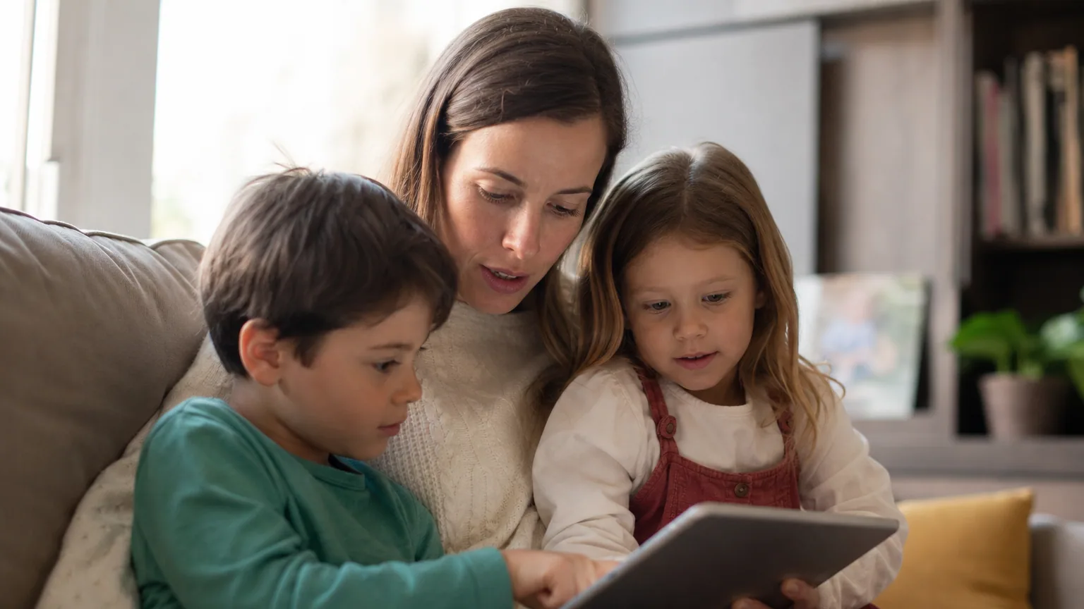 Une mère et ses deux enfants regardent ensemble une tablette sur un canapé dans un salon lumineux.